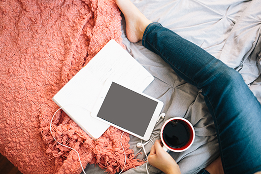 Woman in jeans sitting on bed with coffee, ipad, laptop, earphones